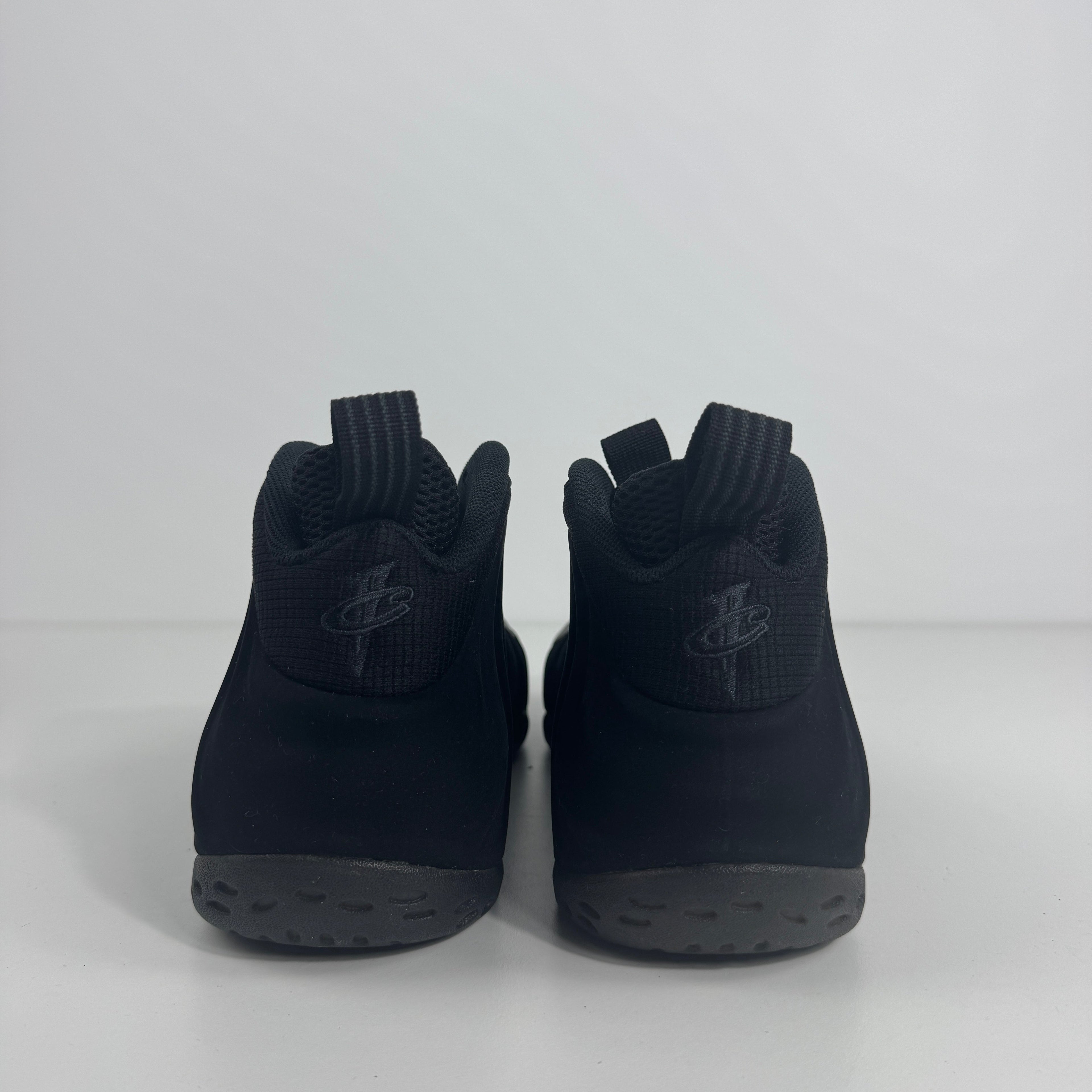 Black NIKE toddler trainers with textured grey soles and rear pull tabs, shown from the back on a white background.