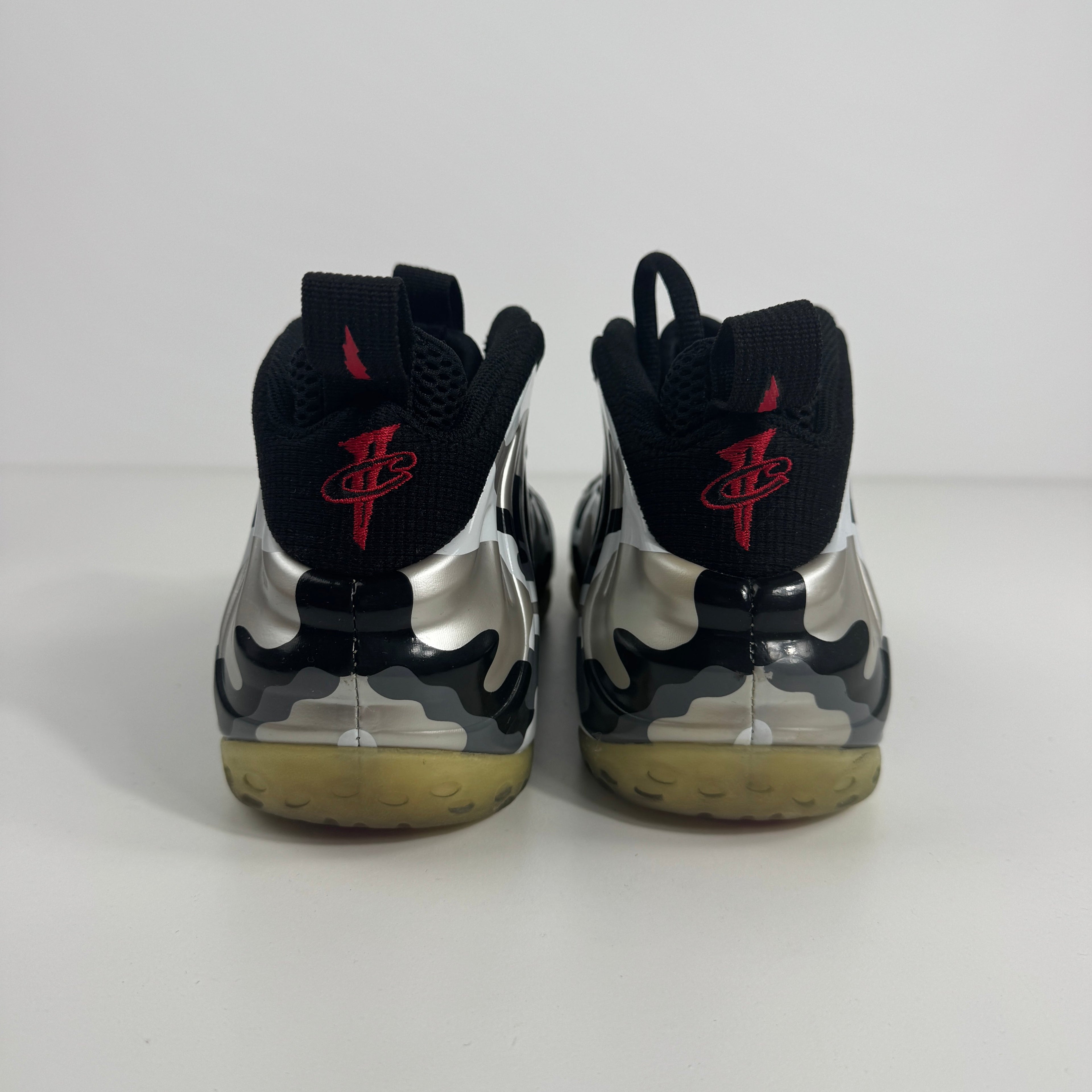 NIKE Air Foamposite One trainers viewed from the back, with a shiny silver and black design, red logo pull tabs and yellowed soles.