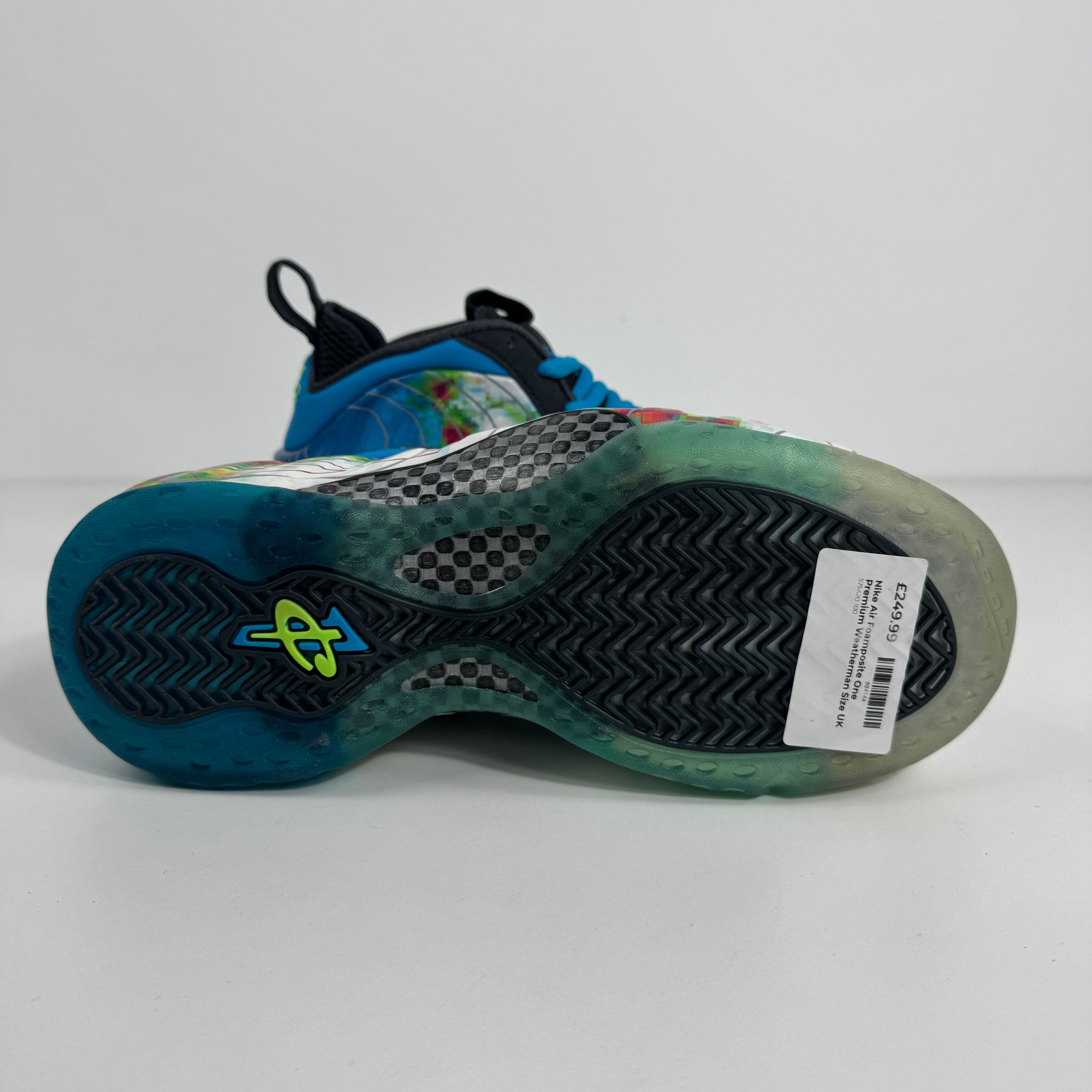 Sole of a Nike Air Foamposite One trainer in blue, green and black with a barcode sticker and yellow logo by the heel on white background.