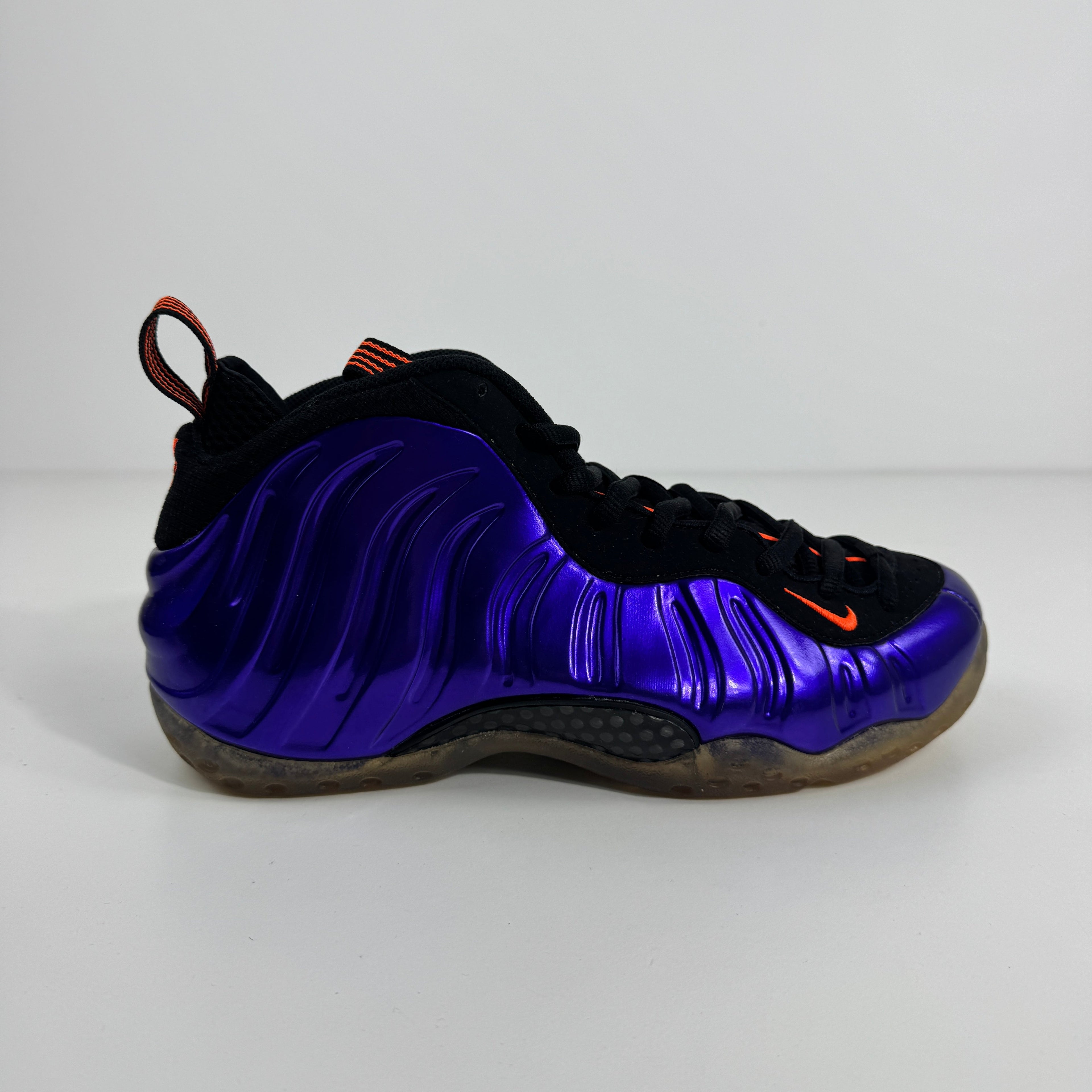 Nike Air Foamposite One trainer with a purple upper, black and orange accents, wavy glossy sides, and black laces.