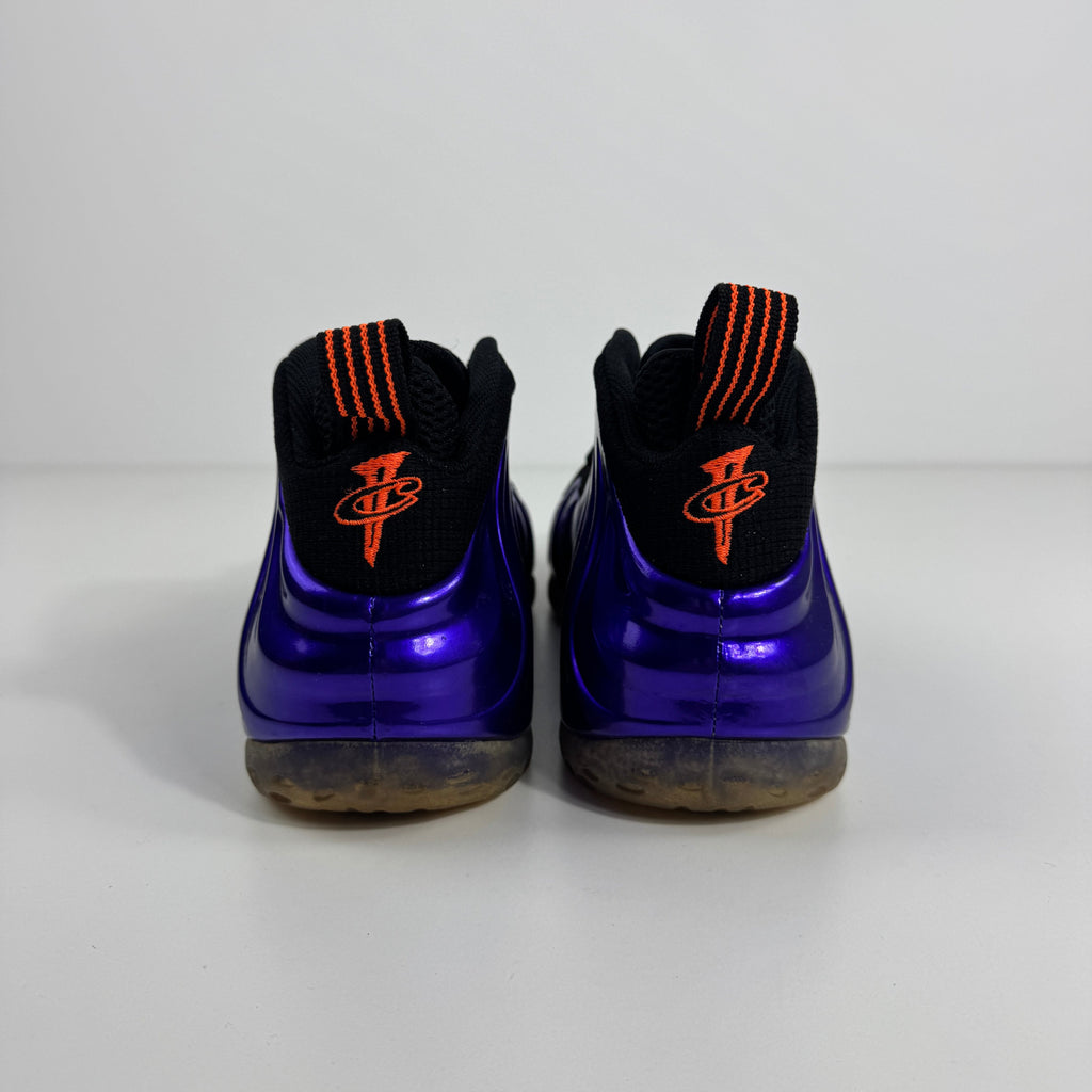 Purple NIKE Air Foamposite One trainers shown from the back with black collars, orange-striped pull tabs and orange heel logos.