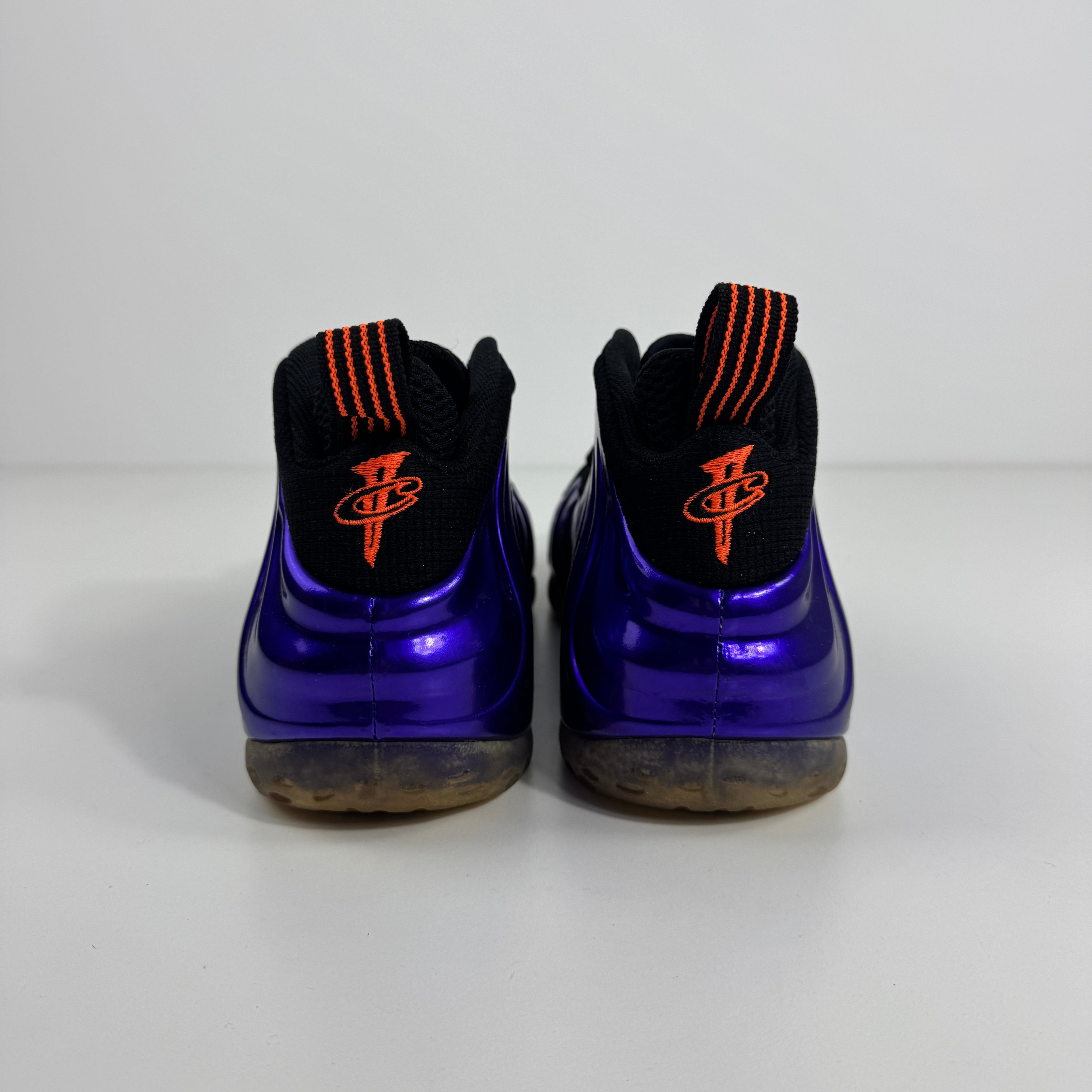 Purple NIKE Air Foamposite One trainers shown from the back with black collars, orange-striped pull tabs and orange heel logos.