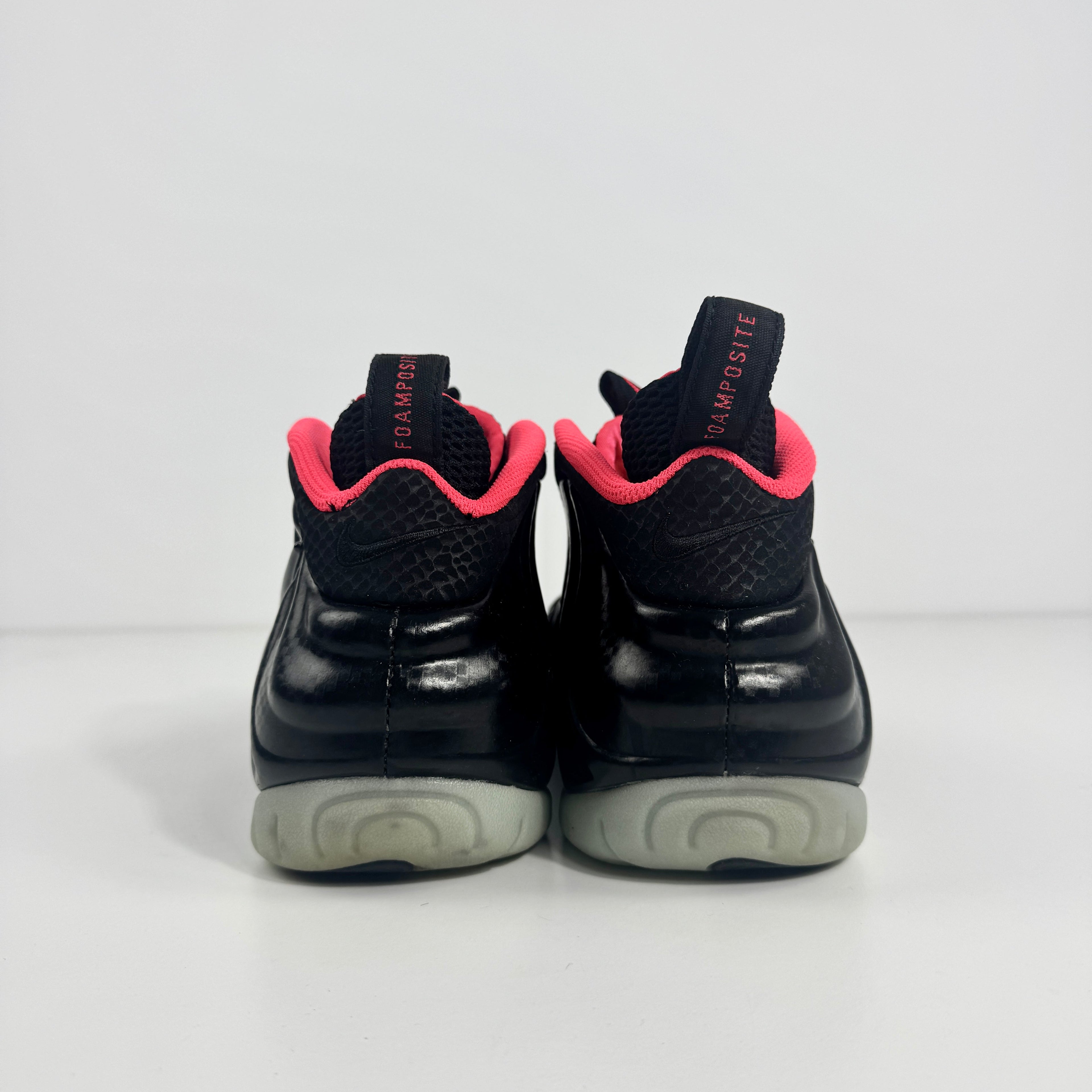 Back view of black Nike trainers with a pink inner lining, grey soles, and black pull tabs featuring red text, on a white background.