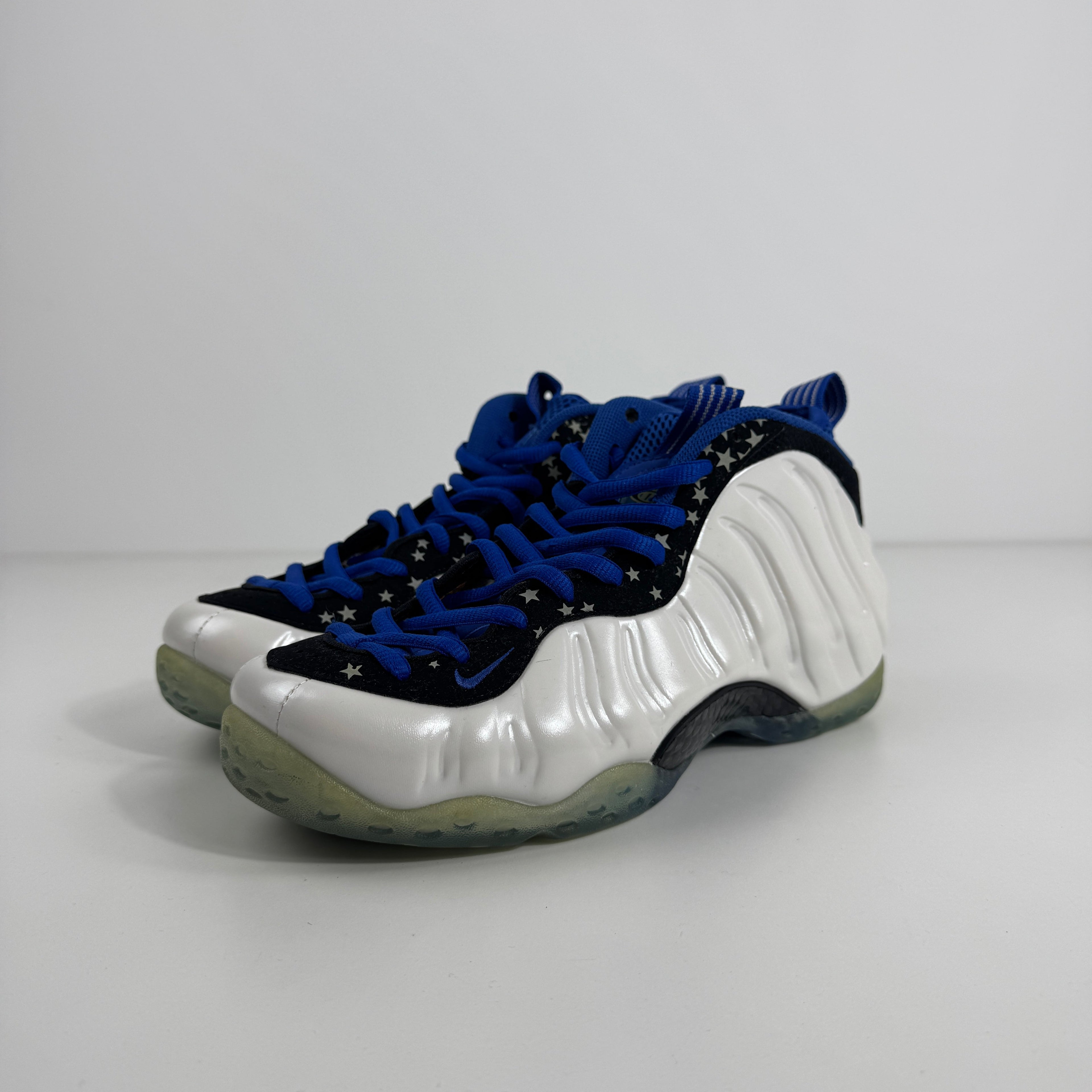 NIKE Air Foamposite Pro trainers with a white wavy shell, blue laces, black uppers with silver stars, and translucent soles on white.