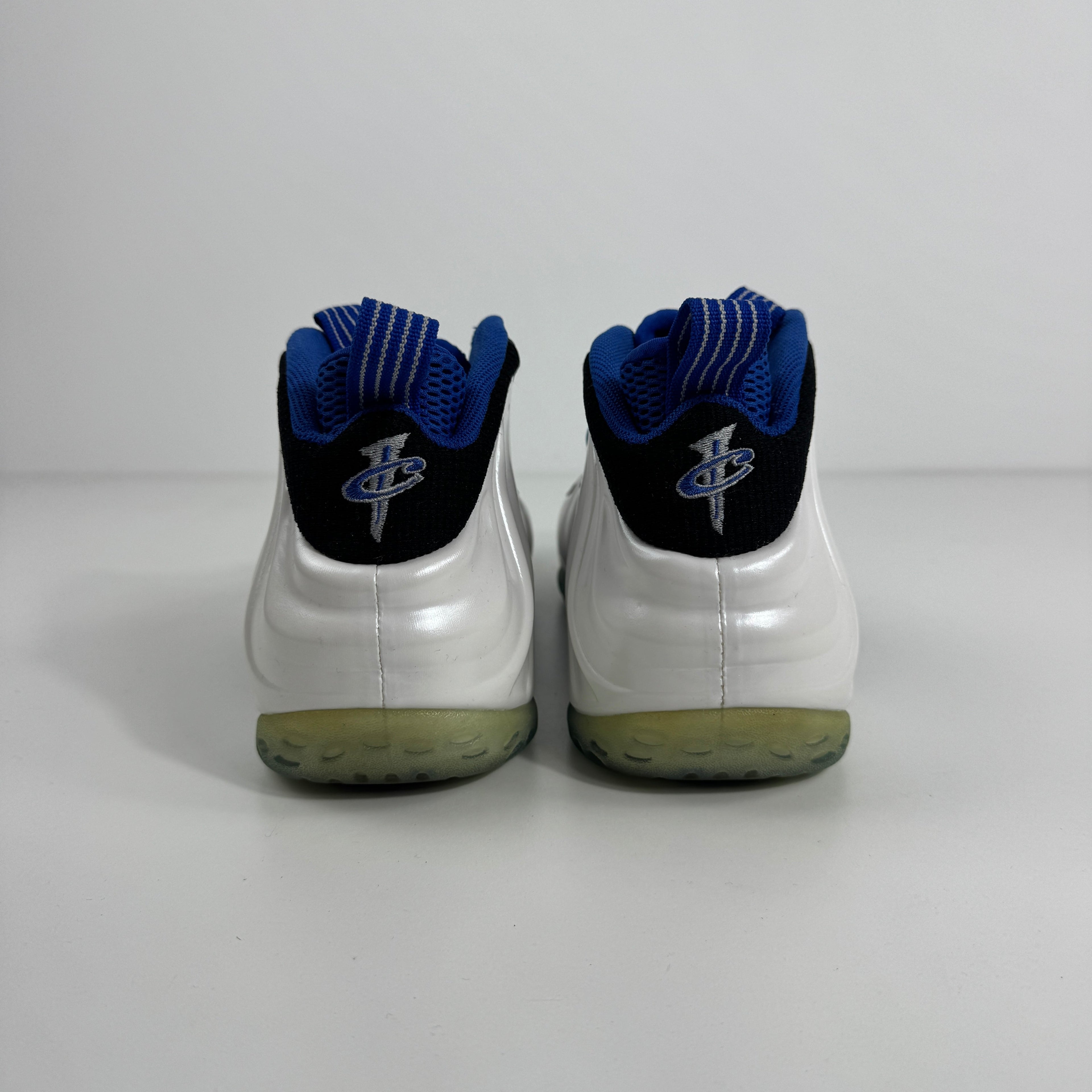 Rear view of white Nike Air Foamposite Pro trainers with translucent soles, blue lining, black heels and silver-blue logo, on white background.