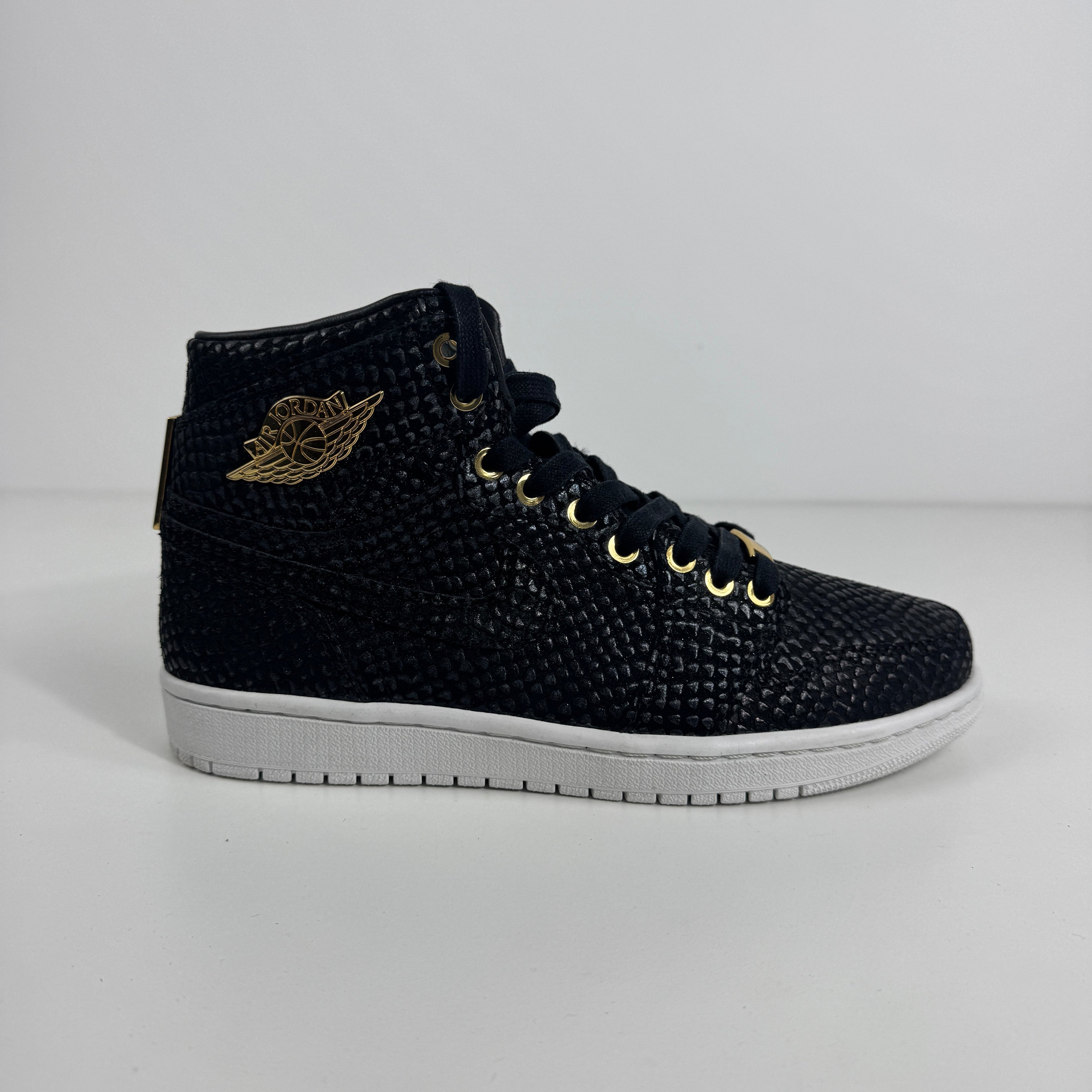 Black Nike high-top trainer with snakeskin texture, gold eyelets and wing logo, and a white rubber sole on a white background.