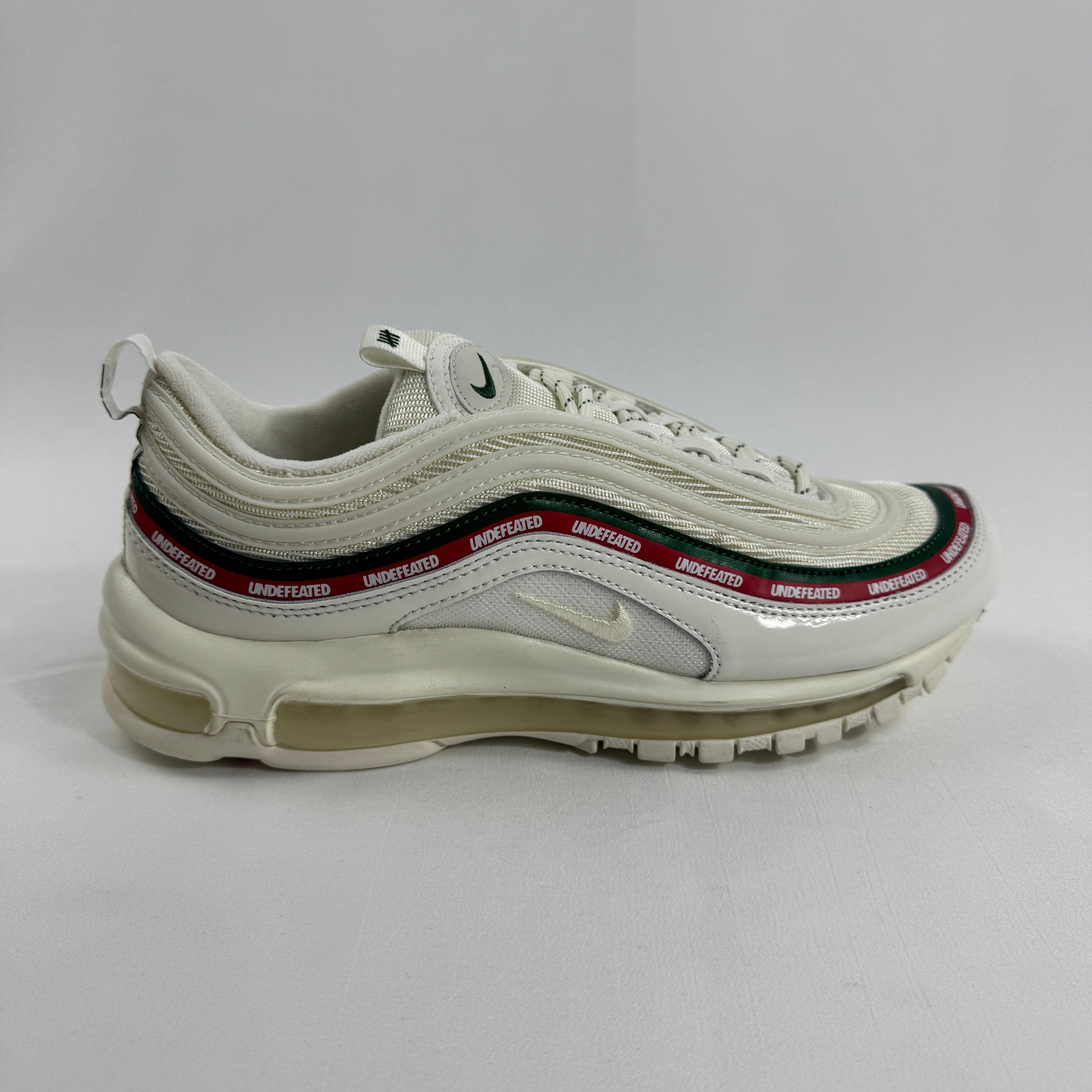 Nike Air Max 97 Undefeated Sail trainers with visible Air cushioning, green and red details, and UNDEFEATED on a red stripe.