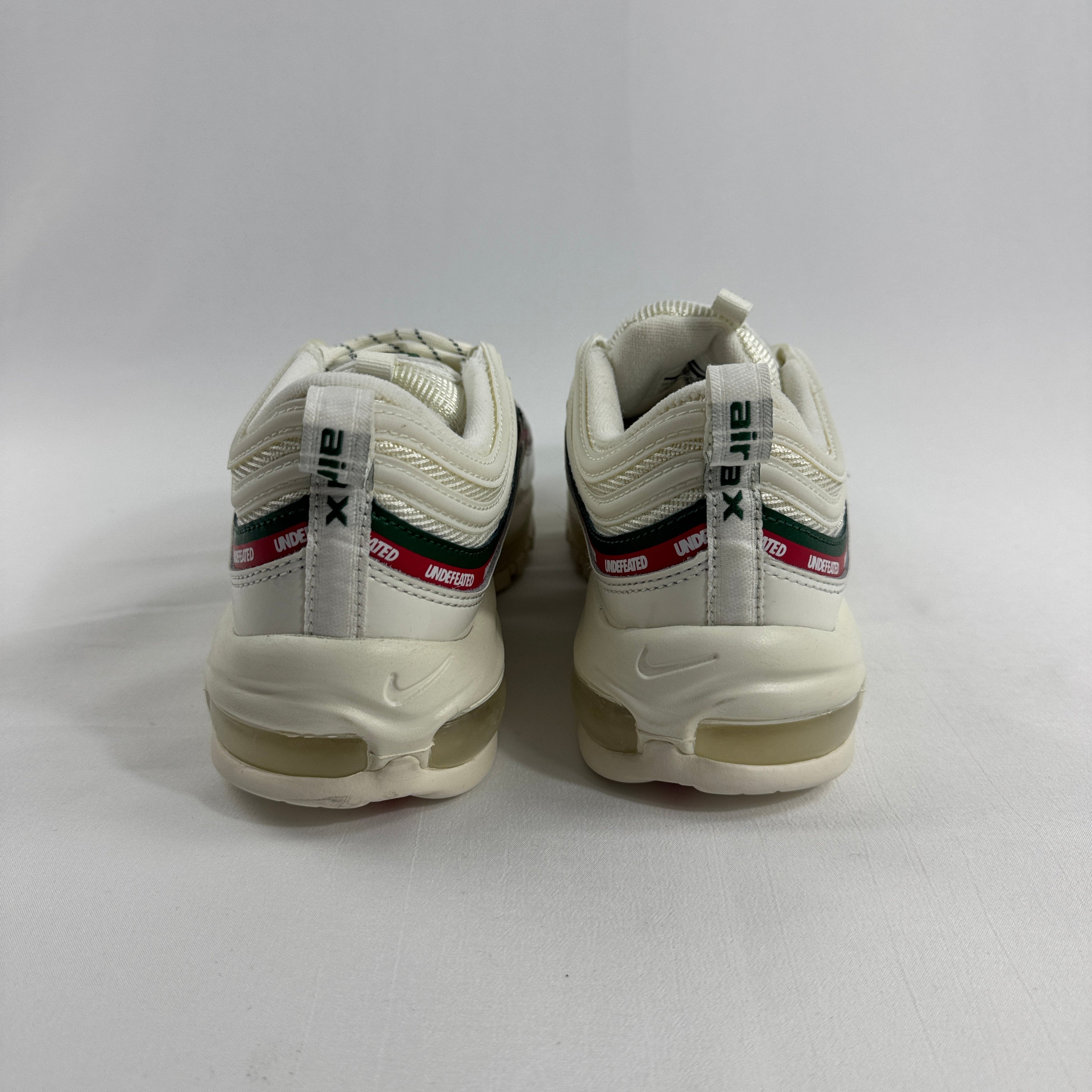 NIKE Air Max 97 Undefeated Sail trainers with white uppers, green and red stripes, and Undefeated detail on the heel, rear view.