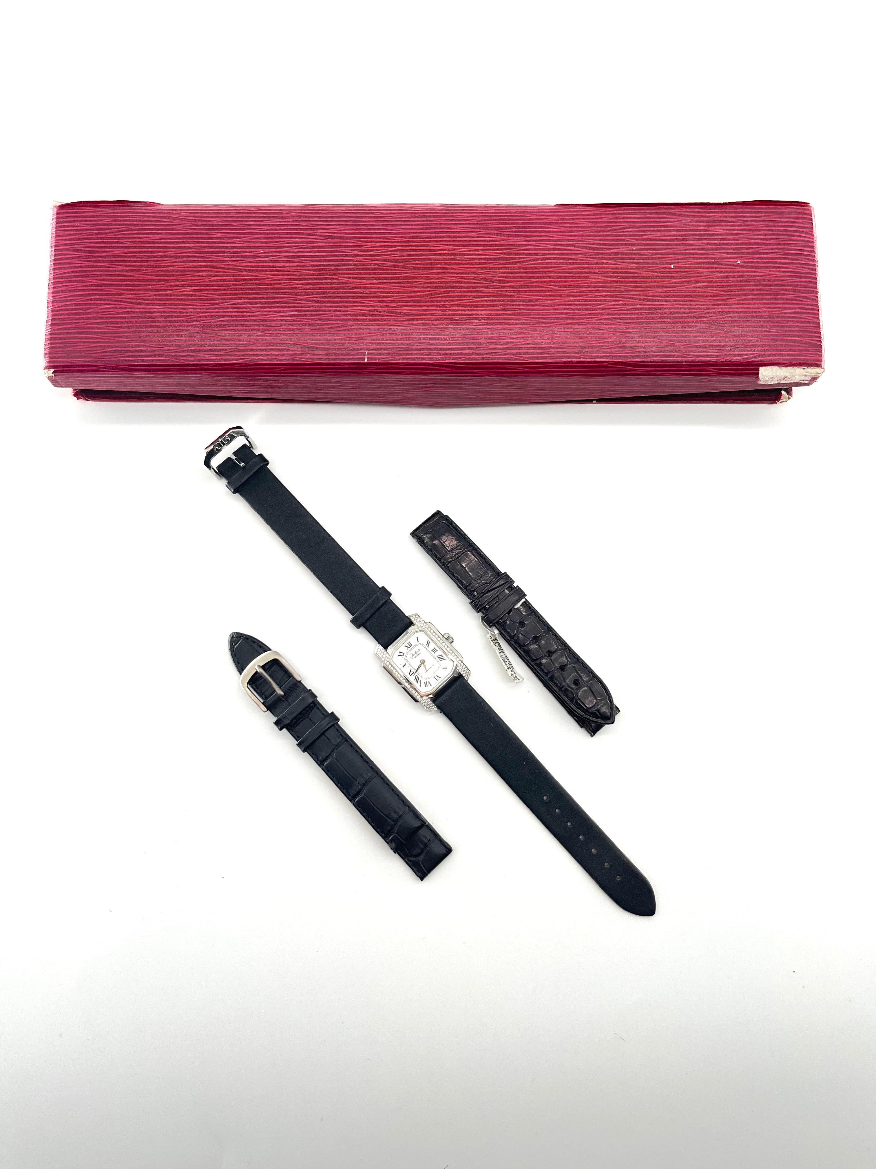A red watch box sits above a silver Glashutte watch with a black strap, shown with two extra straps on a white background.