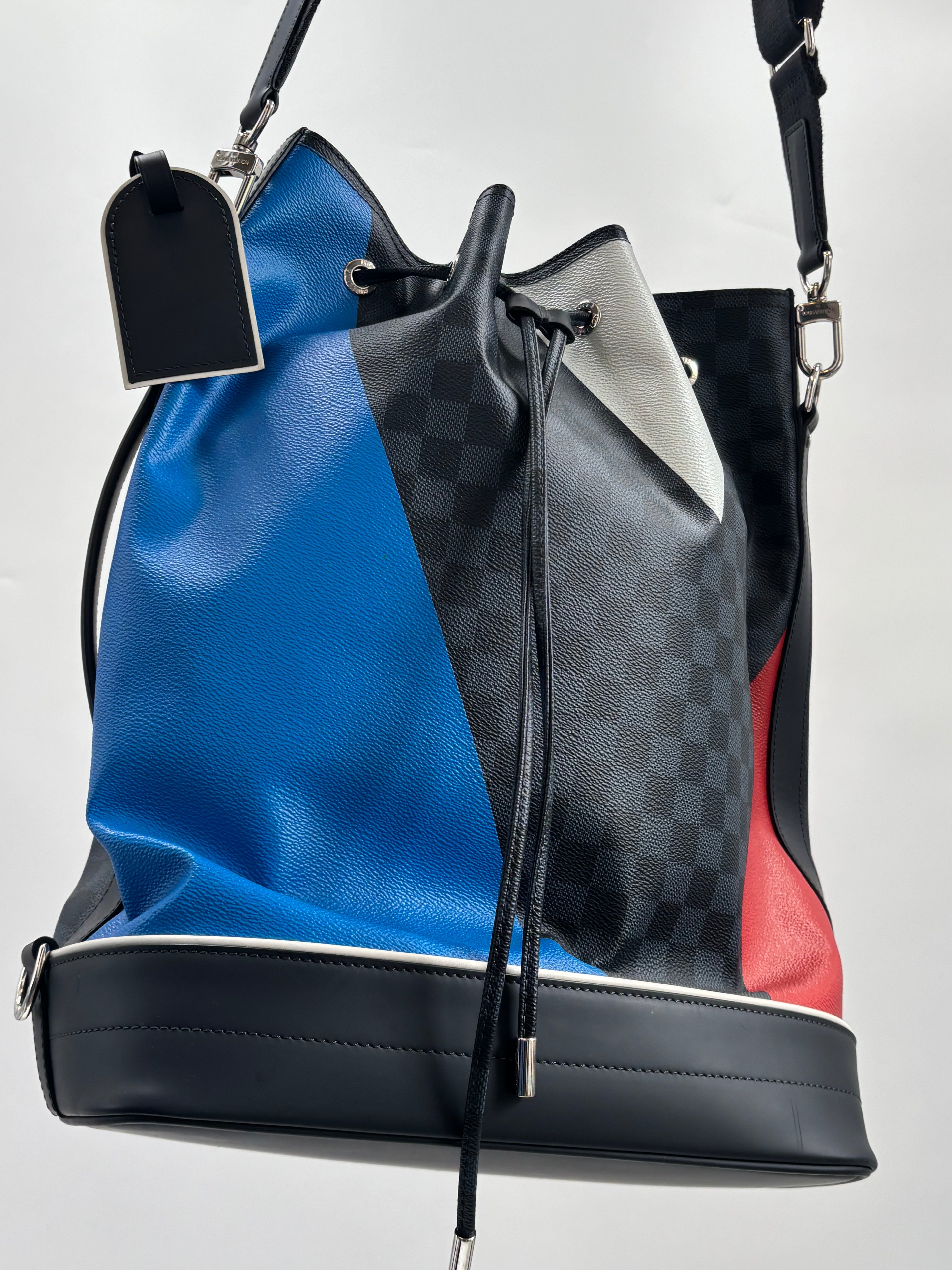 Louis Vuitton bucket bag with blue, red, black and grey check panels, black strap, silver hardware and drawstring on white background.
