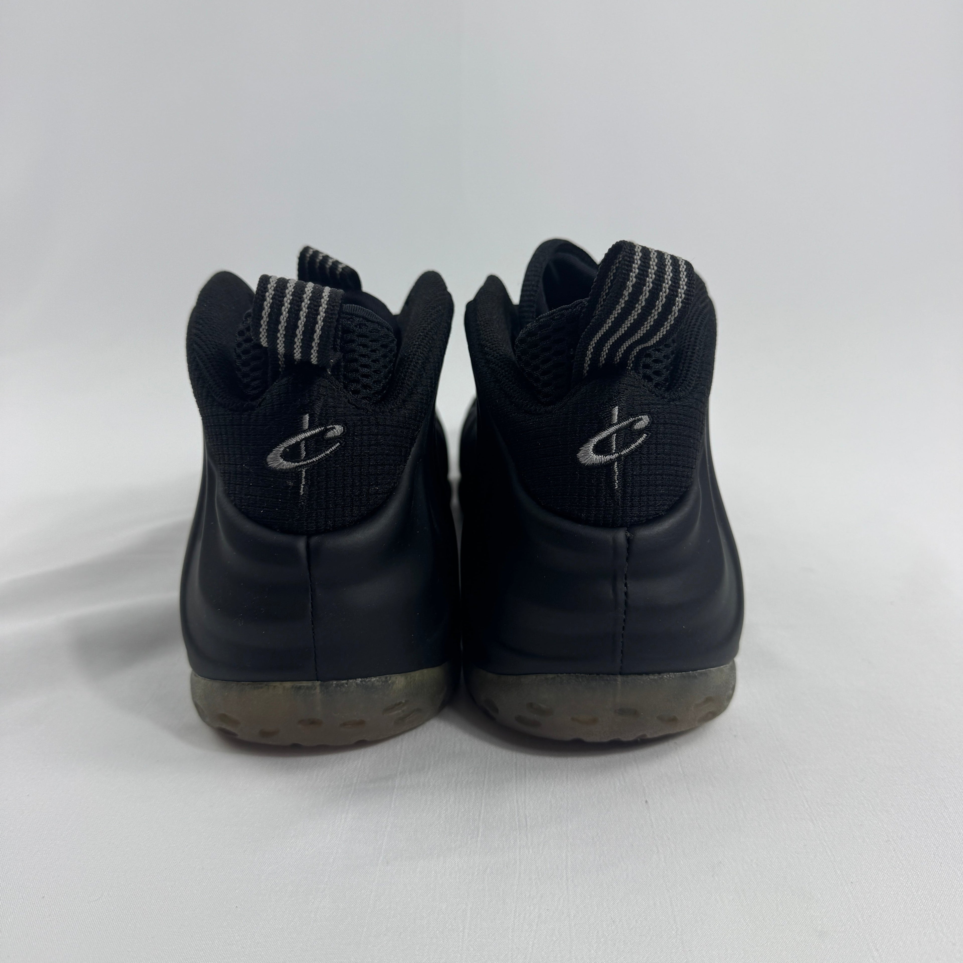 Nike Air Foamposite One trainers in black with striped pull tabs, stylised heel logo and semi-translucent midsoles on white background.