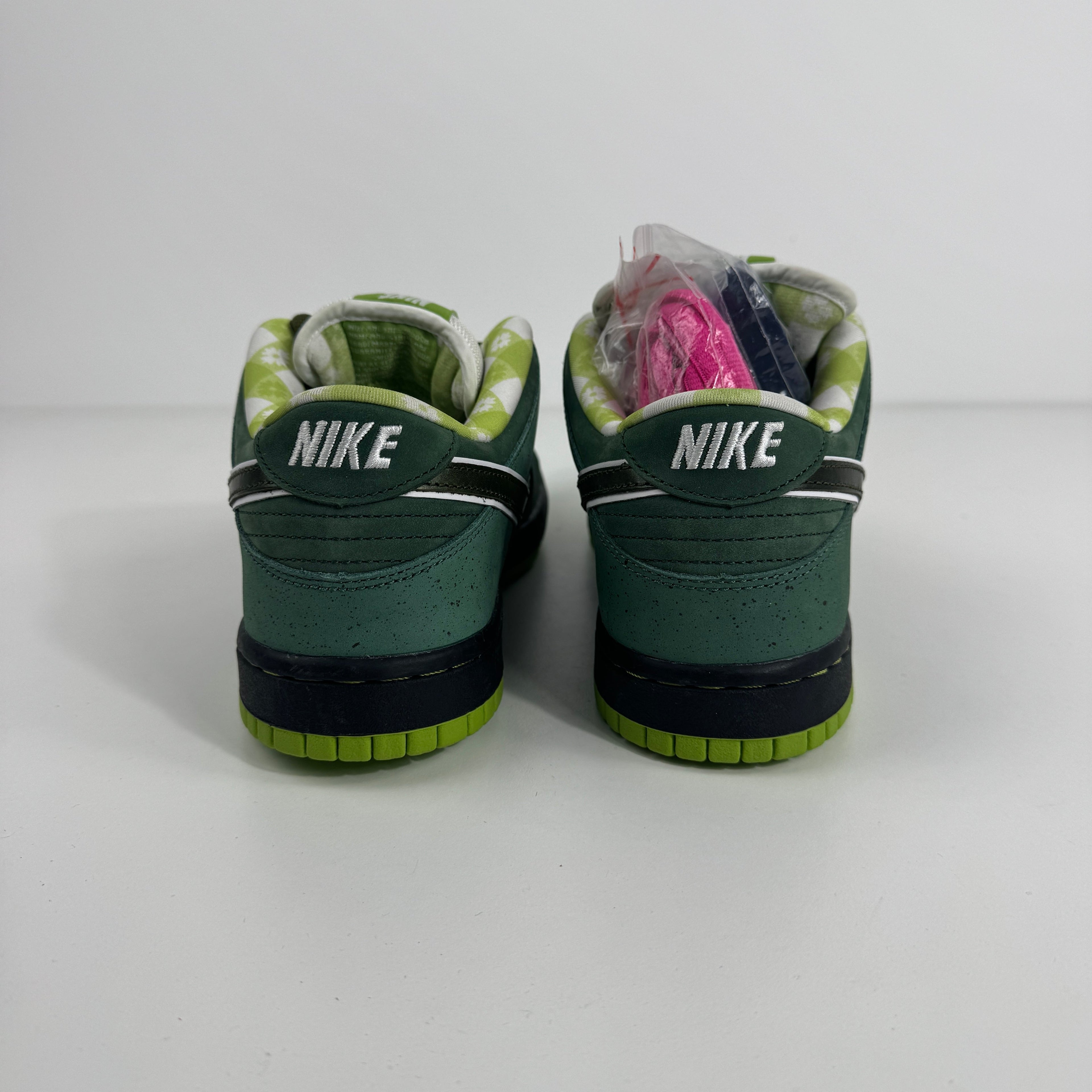 Green Nike SB Dunk Low sneakers viewed from the back, with a checkered lining and extra pink and purple laces on the left shoe.