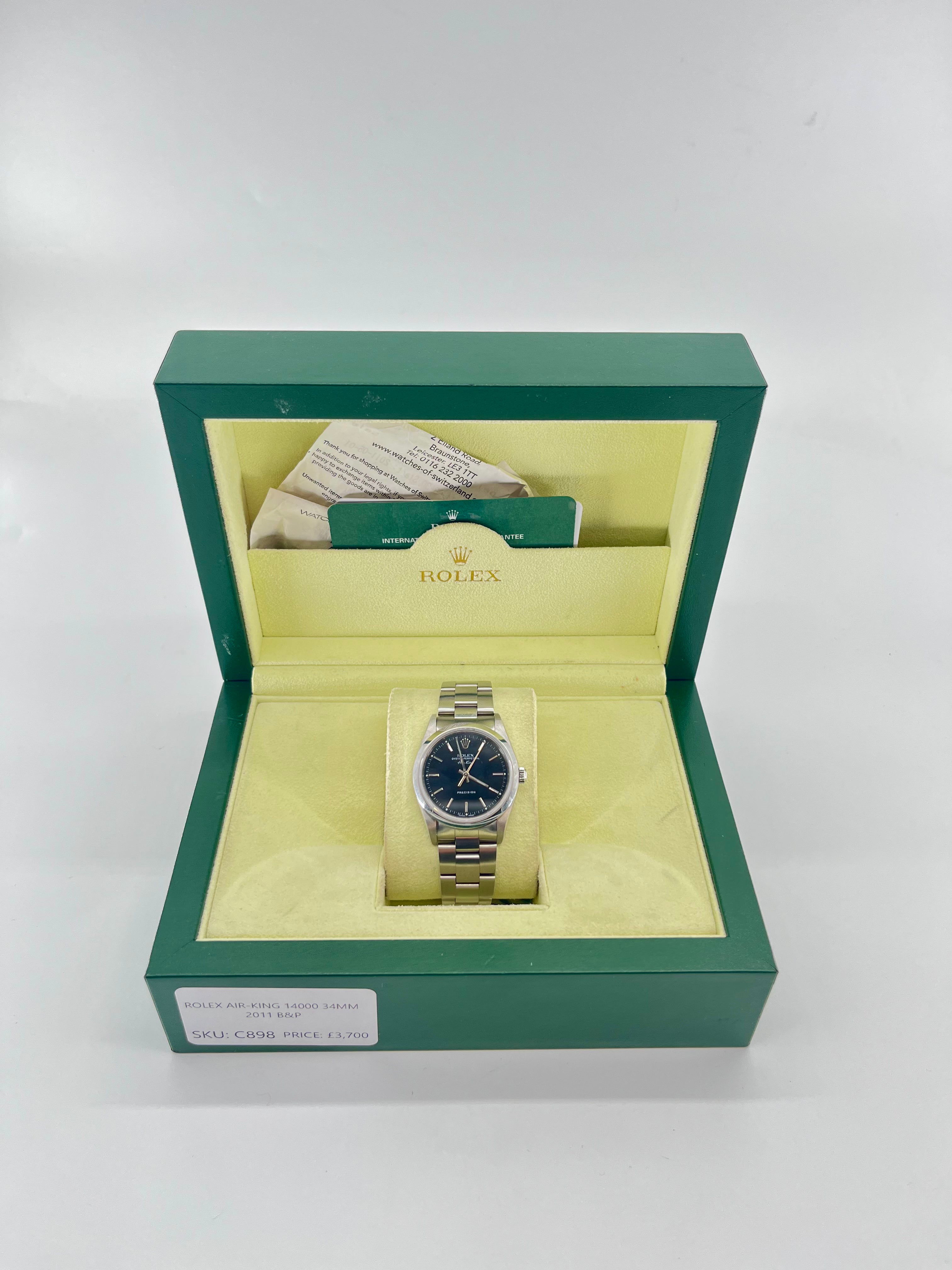 A silver Rolex watch with a dark dial in an open green and yellow box, shown with papers, card, and a price tag behind it.