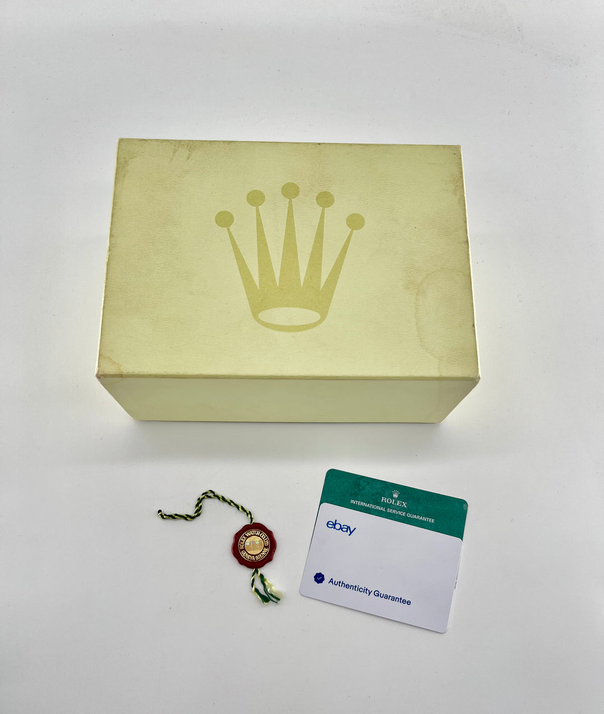 A beige Rolex box with a crown logo, shown on a white surface with a red tag, green and white string, and an eBay card.