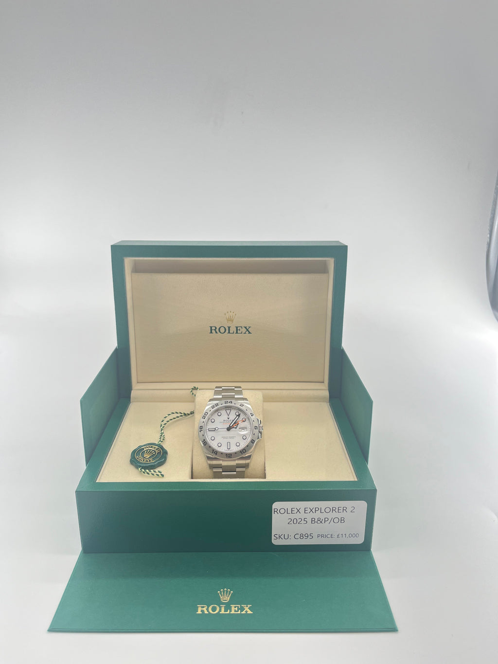A Rolex Explorer 2 watch with a white dial and orange hand displayed in an open green and beige box, next to a green tag and label.