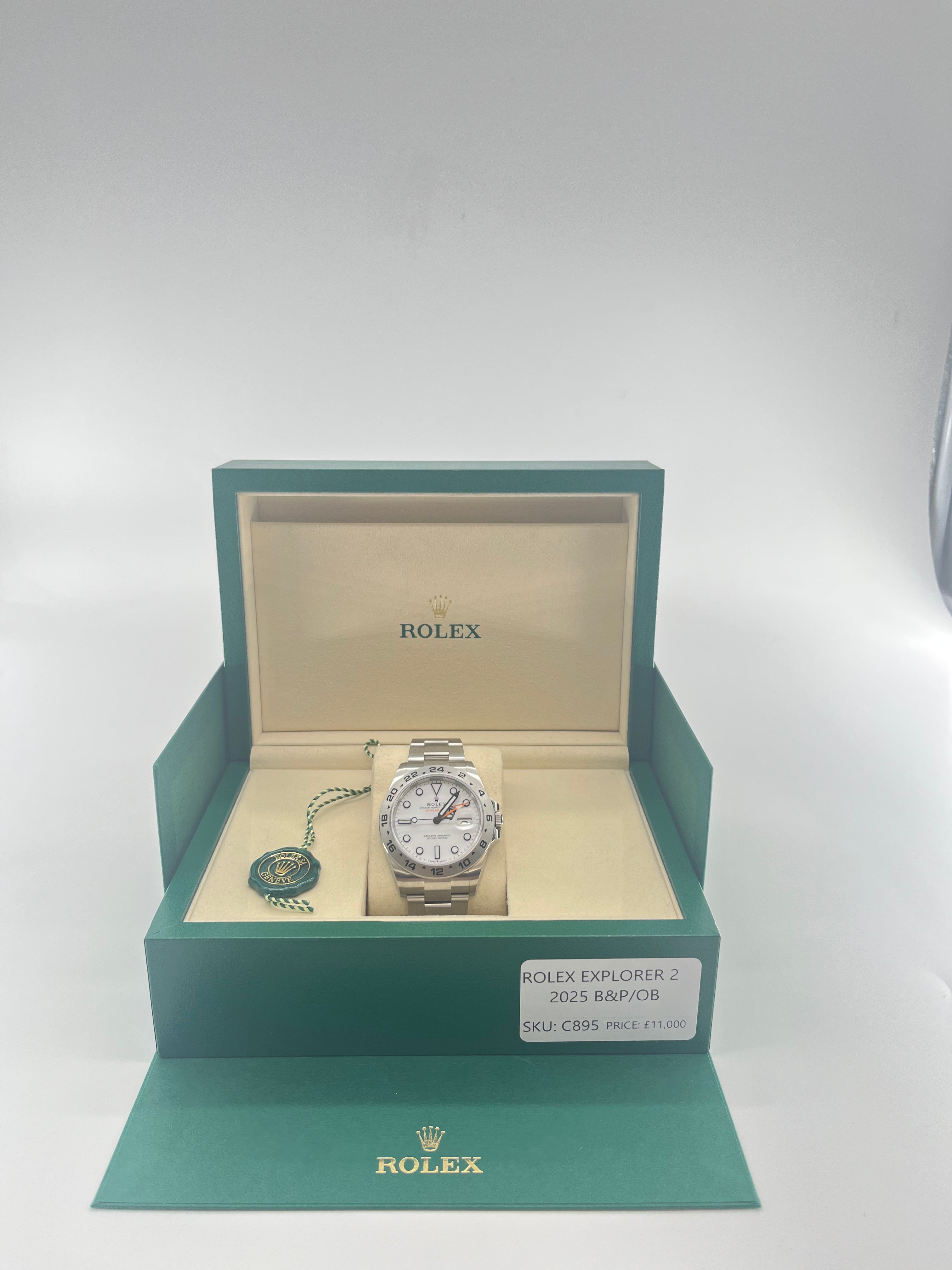 A Rolex Explorer 2 watch with a white dial and orange hand displayed in an open green and beige box, next to a green tag and label.