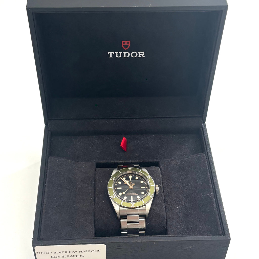 A TUDOR wristwatch with a green bezel and black dial rests on a cushion in a branded box, shown with its attached tag.