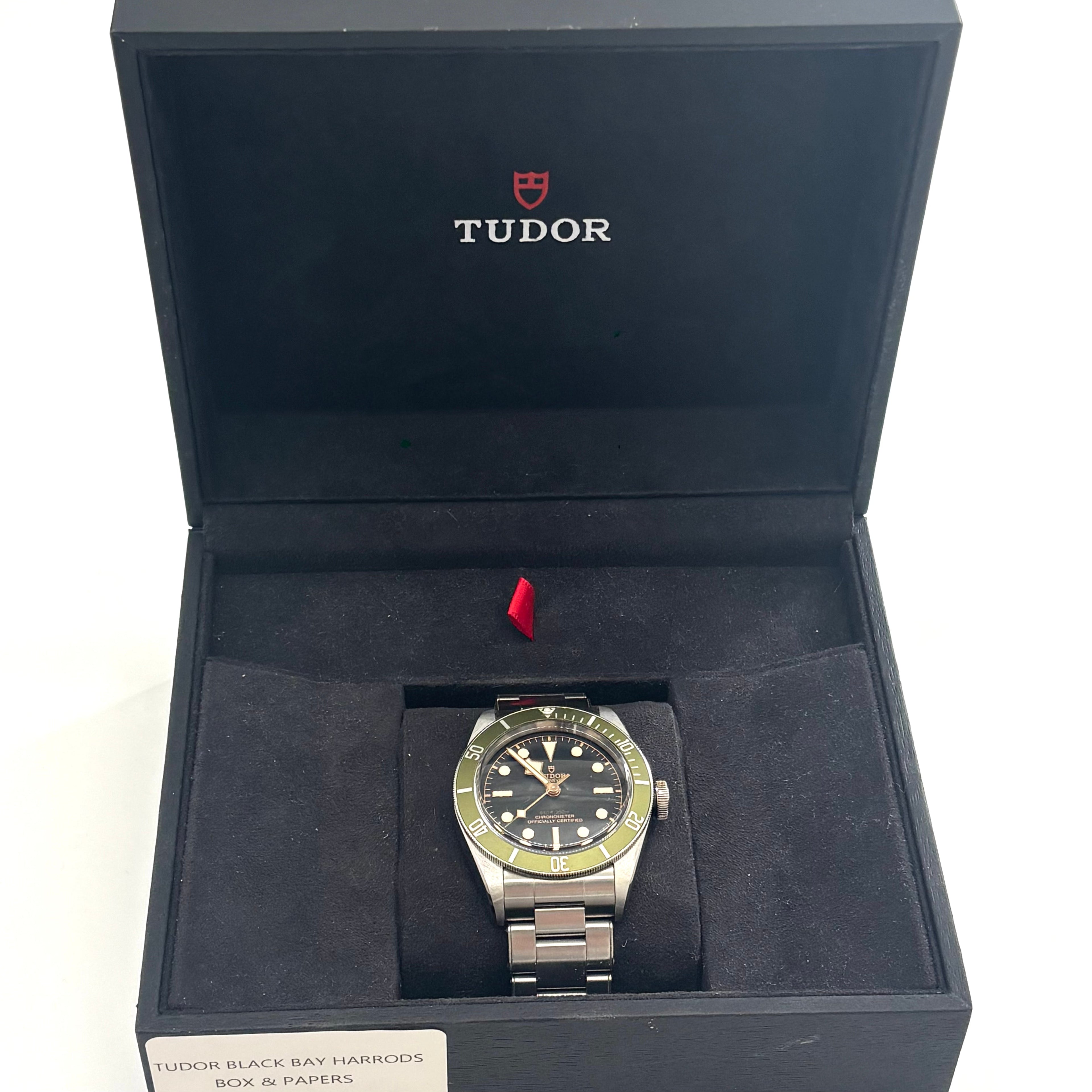 A TUDOR wristwatch with a green bezel and black dial rests on a cushion in a branded box, shown with its attached tag.