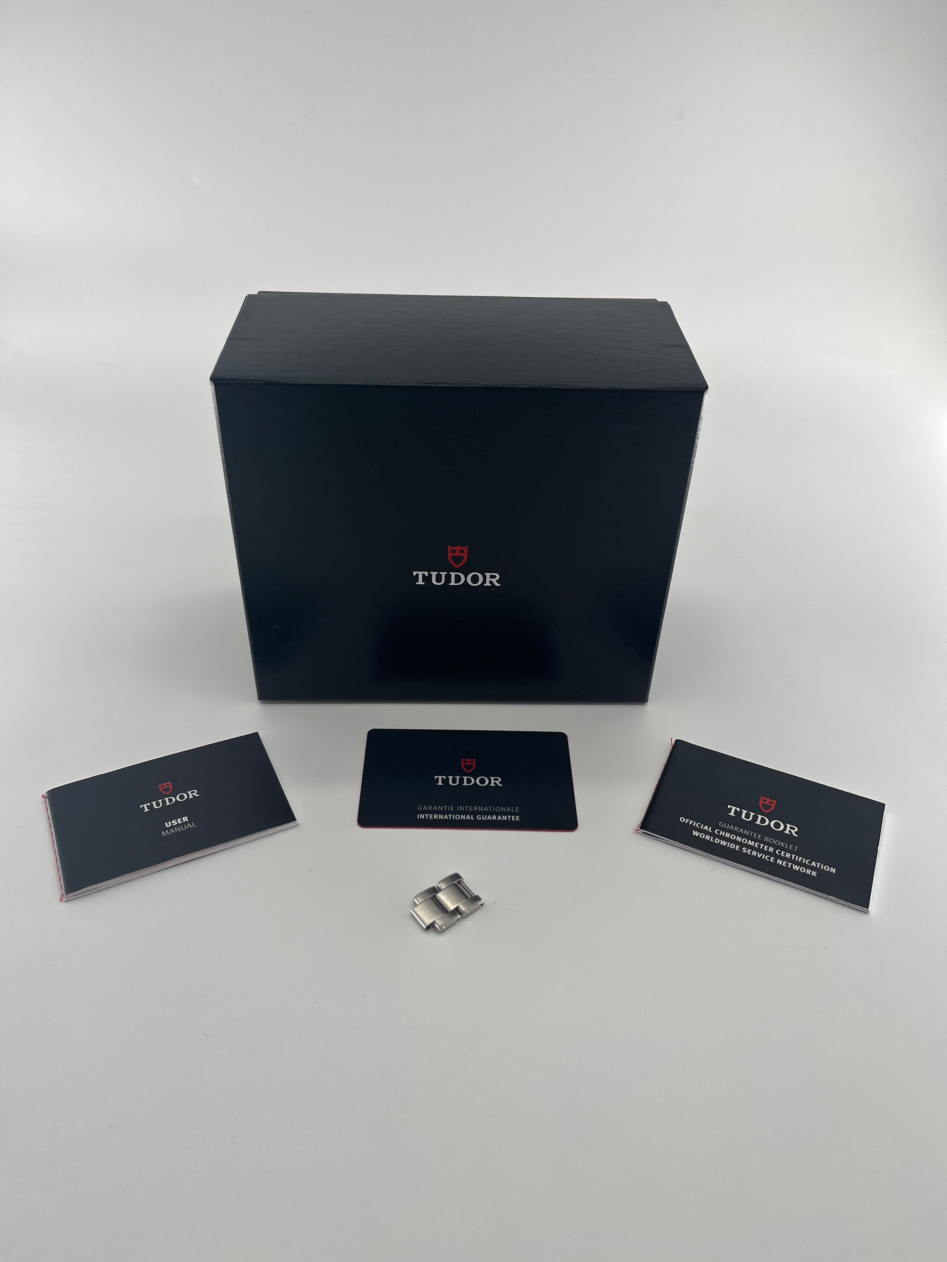 A black Tudor presentation box is centred with a booklet, guarantee card, certificate, and two metal links arranged in front.