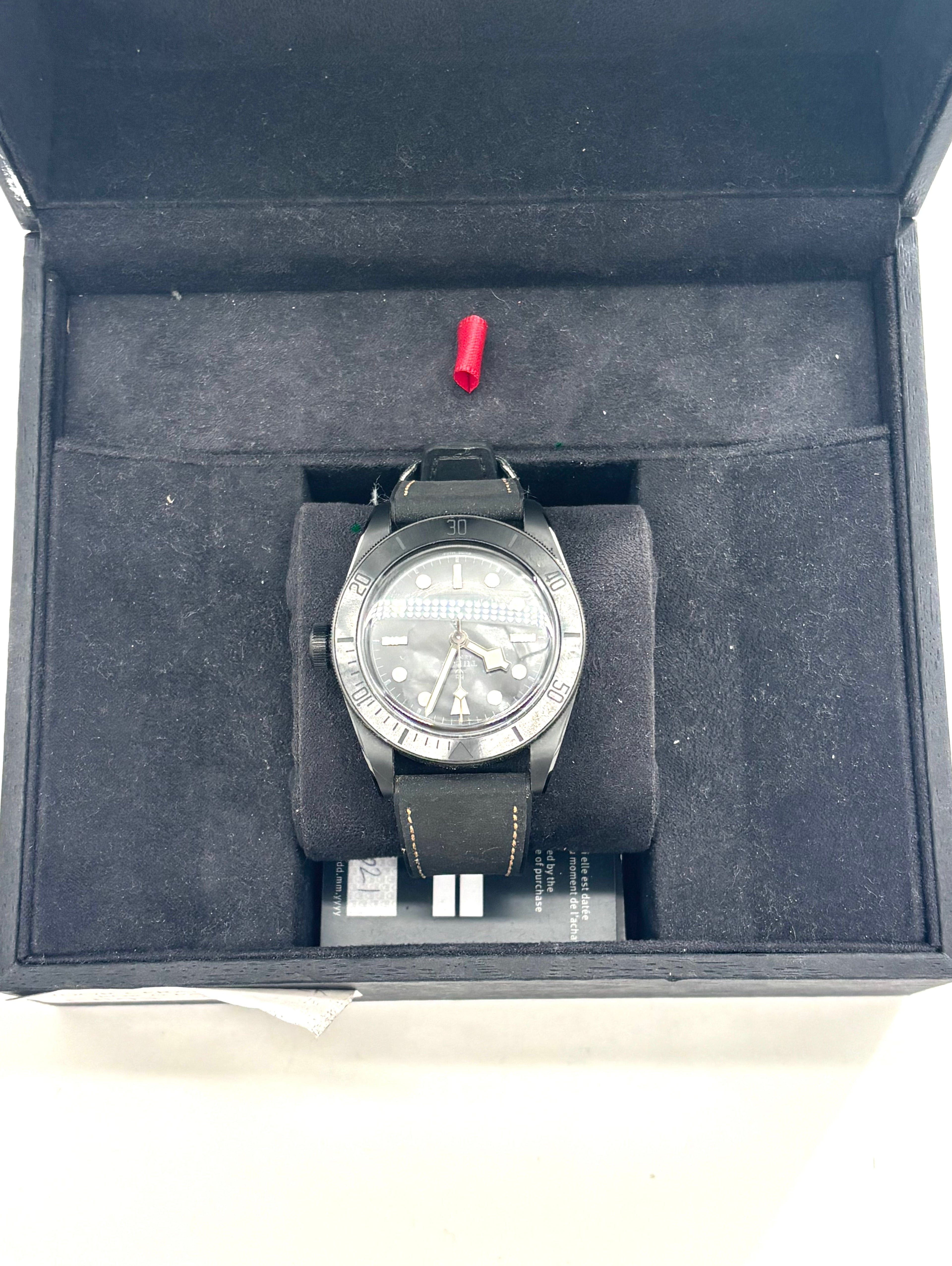 A TUDOR black ceramic wristwatch with a leather strap rests in a velvet-lined box, with a small red detail and card beside it.