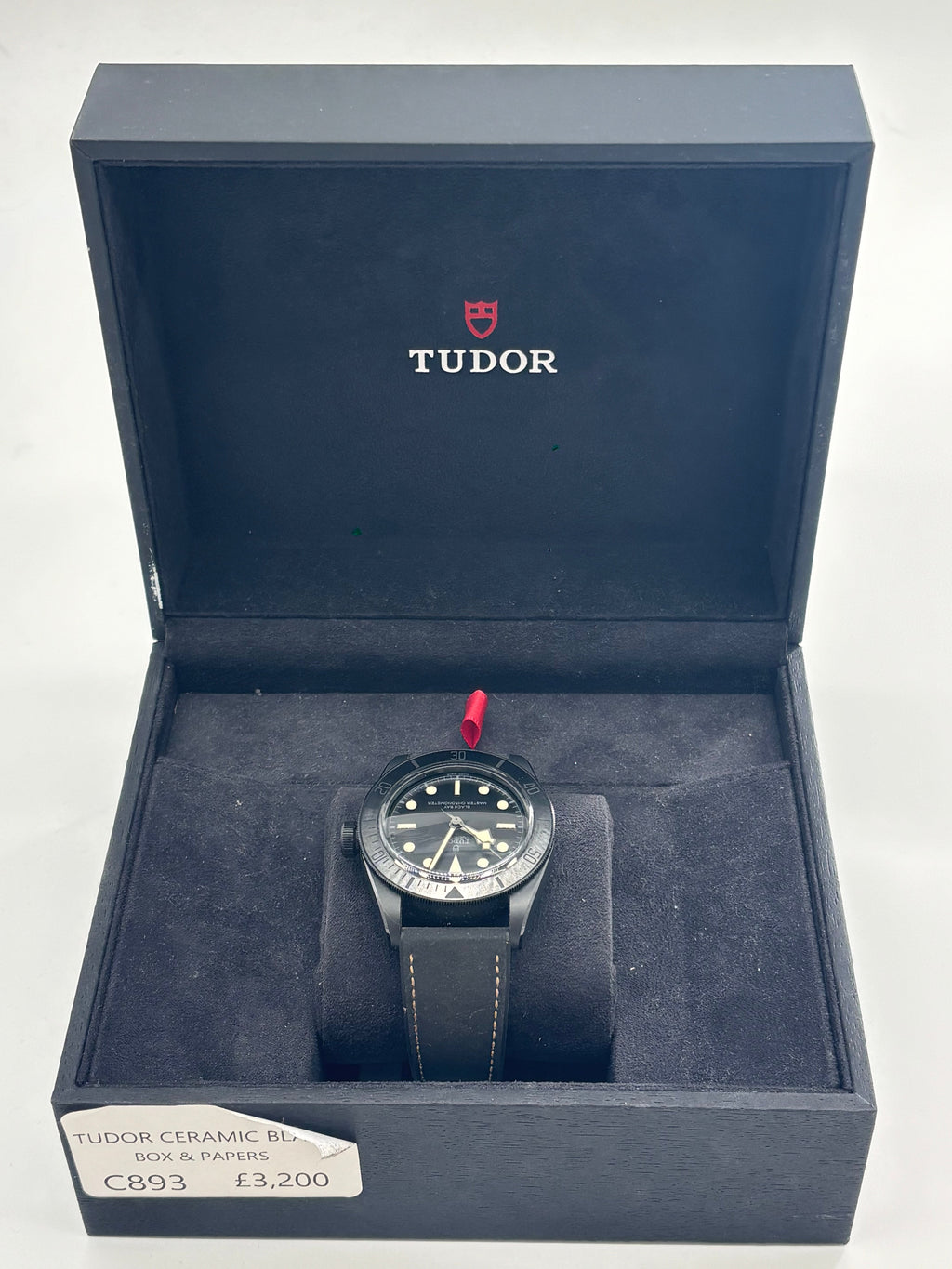 A TUDOR black ceramic automatic watch with a black strap stands upright in an open box, label and logo visible inside the lid.