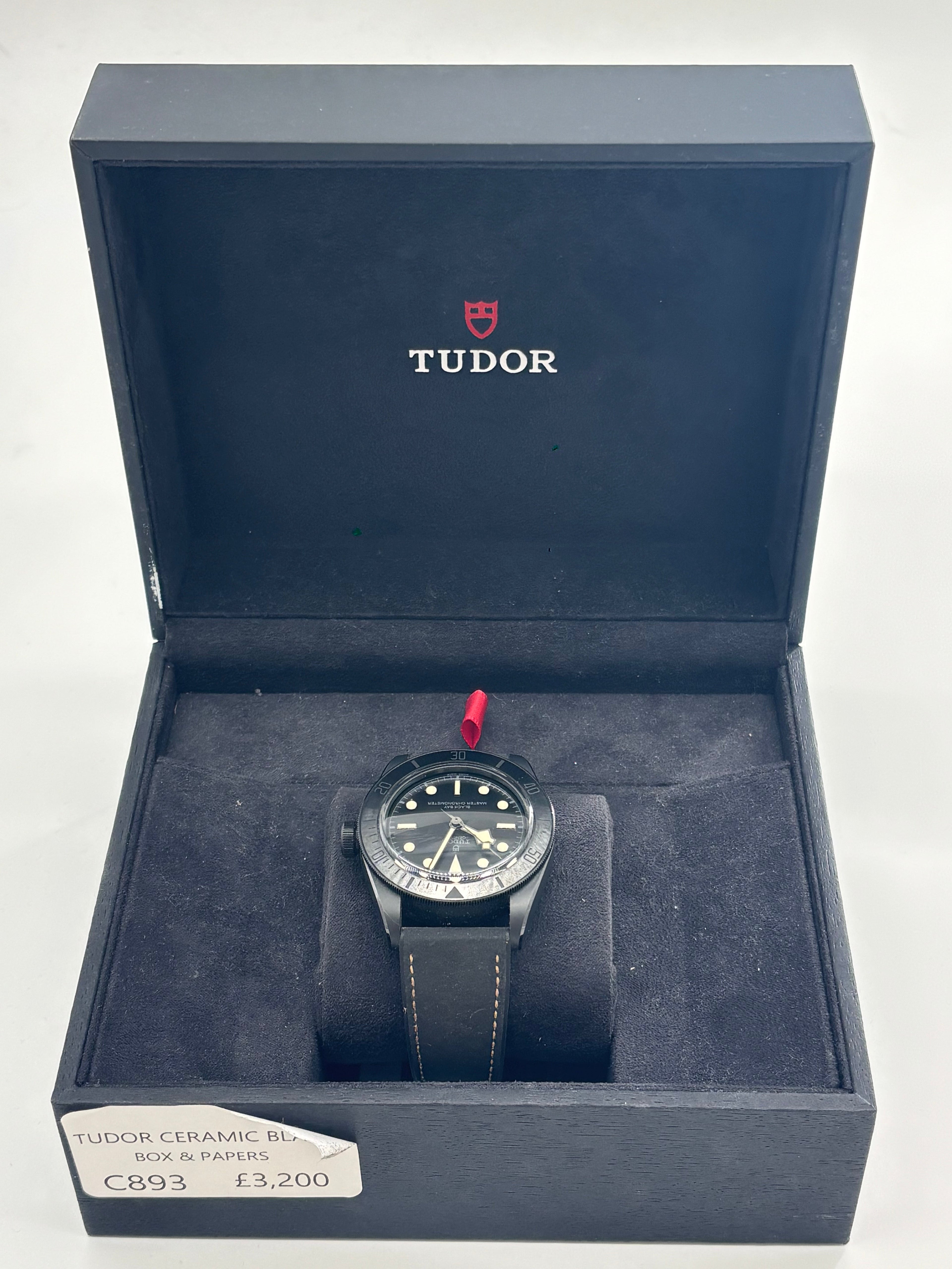 A TUDOR black ceramic automatic watch with a black strap stands upright in an open box, label and logo visible inside the lid.