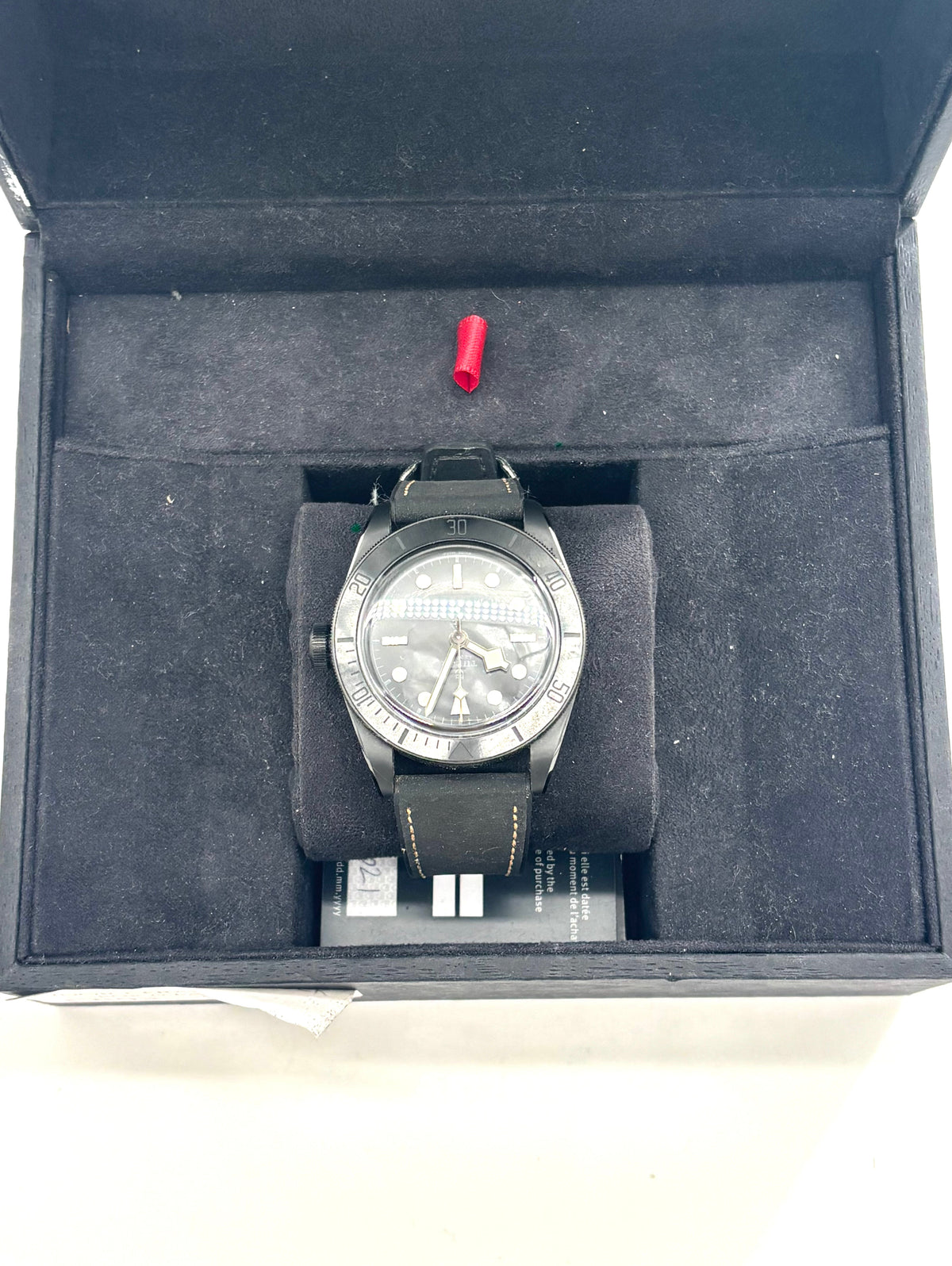 A TUDOR black ceramic wristwatch with a leather strap rests in a velvet-lined box, with a small red detail and card beside it.